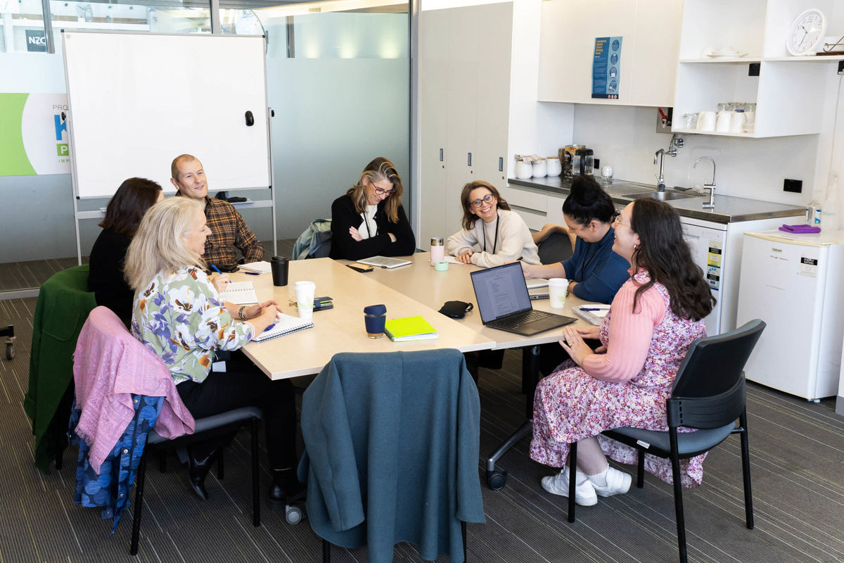 Group meeting around a table in the Coastlands Aquatics Centre meeting room that can be hired by community groups. There is a kitchenette and white board in the background.